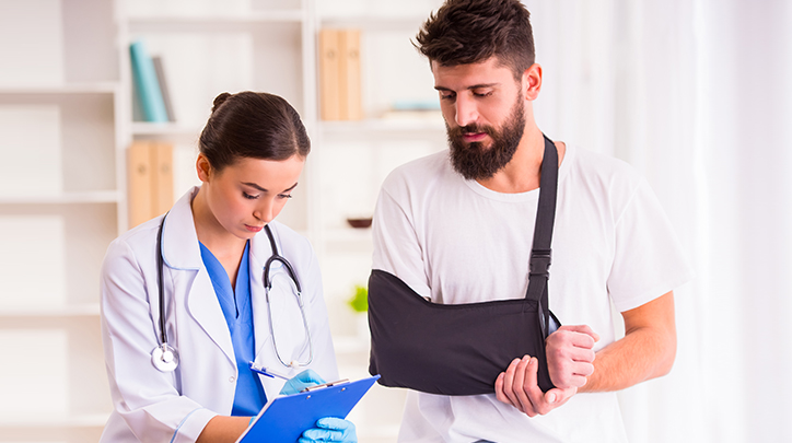 Man in a sling looks over doctor's shoulder, who is writing on a clipboard